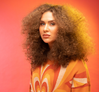 Alex Hosking posing against a warm orange gradient background wearing a retro-inspired patterned outfit with voluminous curly hair.