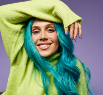 Tigerlily smiling in a lime green oversized jumper against a purple studio backdrop with bright turquoise hair.